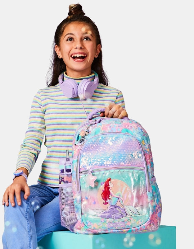 Girl smiling while holding a Smiggle Disney Princess Little Mermaid Classic Backpack, featuring Ariel design and purple headphones around her neck.